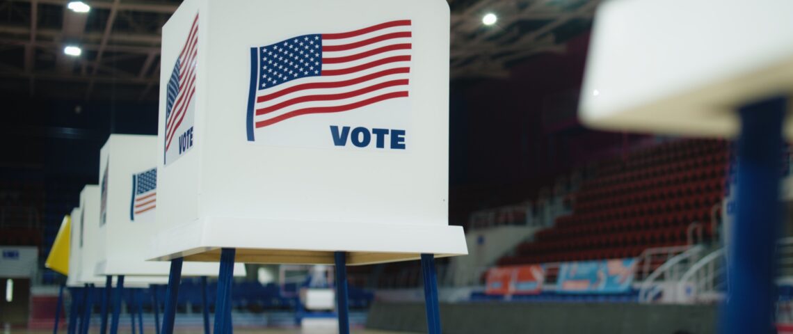 A row of voting booths stands in a hall with the American flag printed on the side and the word “vote”.