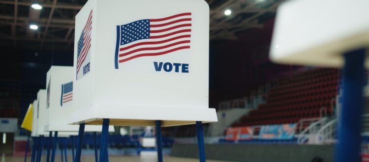 A row of voting booths stands in a hall with the American flag printed on the side and the word “vote”.