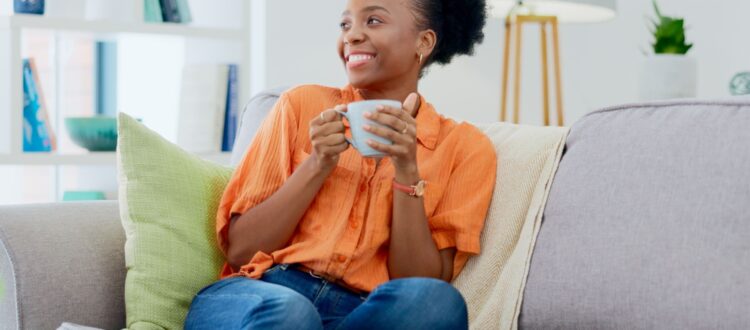 A woman sits on her sofa drinking a cup of tea and smiling.