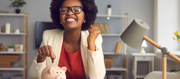 Excited woman sitting at a desk putting a coin into a piggybank