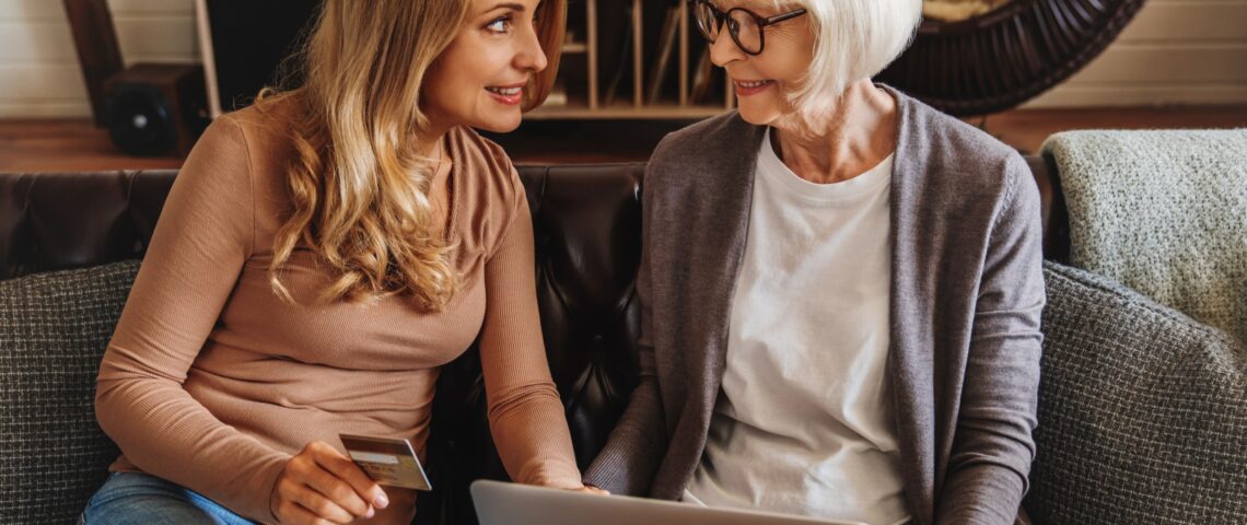 Senior mother holds laptop and smiles at her daughter, who is holding a bank card