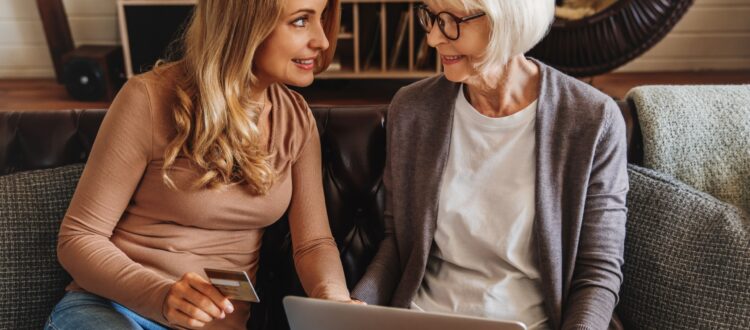 Senior mother holds laptop and smiles at her daughter, who is holding a bank card