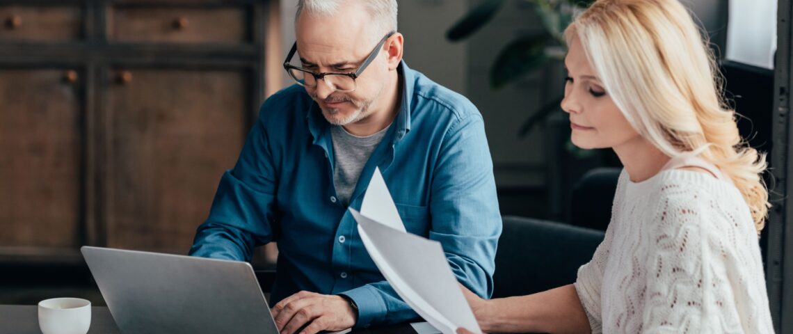 Couple in their 50s look at a laptop and documents, with a calculator and a tablet