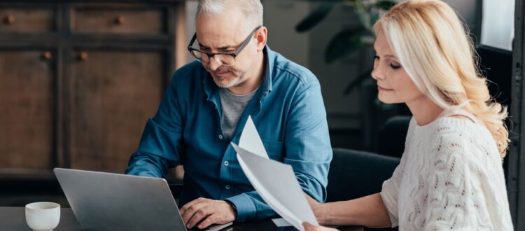 Couple in their 50s look at a laptop and documents, with a calculator and a tablet
