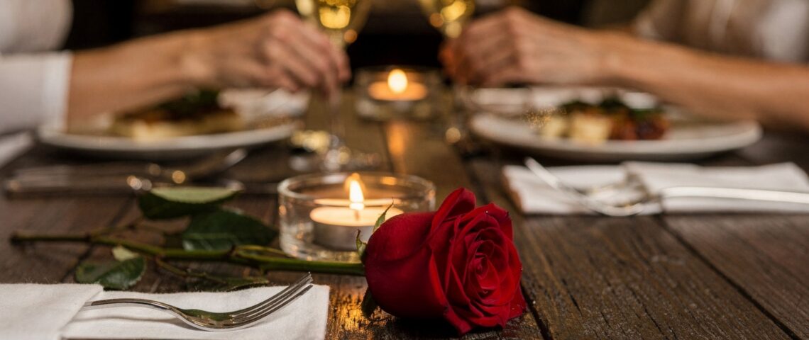 Red rose lying on a dinner table with a couple holding hands in the background.