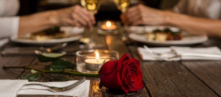 Red rose lying on a dinner table with a couple holding hands in the background.
