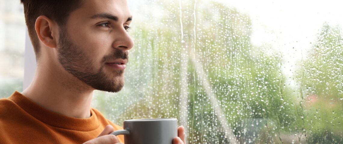 A man looking out at the rain, holding a cup