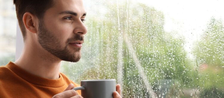 A man looking out at the rain, holding a cup